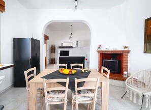 A dining area with a table, chairs, a fruit bowl, a black refrigerator, a fireplace, and a kitchen in the background. - Eden Villa Privata Sul Mare (Porto Cesareo)
