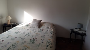 Un dormitorio con colcha estampada, lámpara de noche, mesita de noche y ventana que deja entrar luz natural.