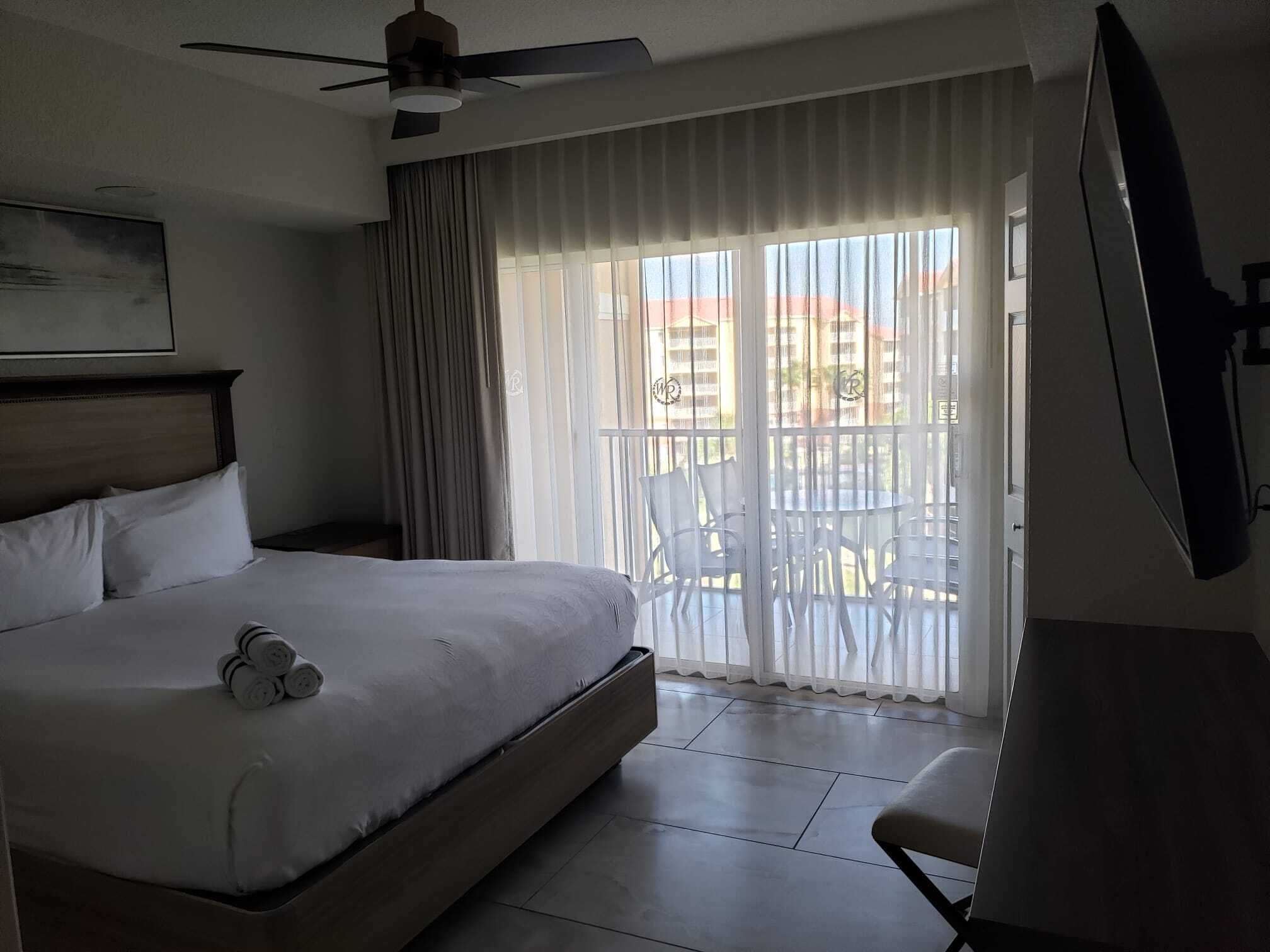 A hotel room with a bed, a ceiling fan, a window with curtains, a balcony with chairs, and a TV mounted on the wall.