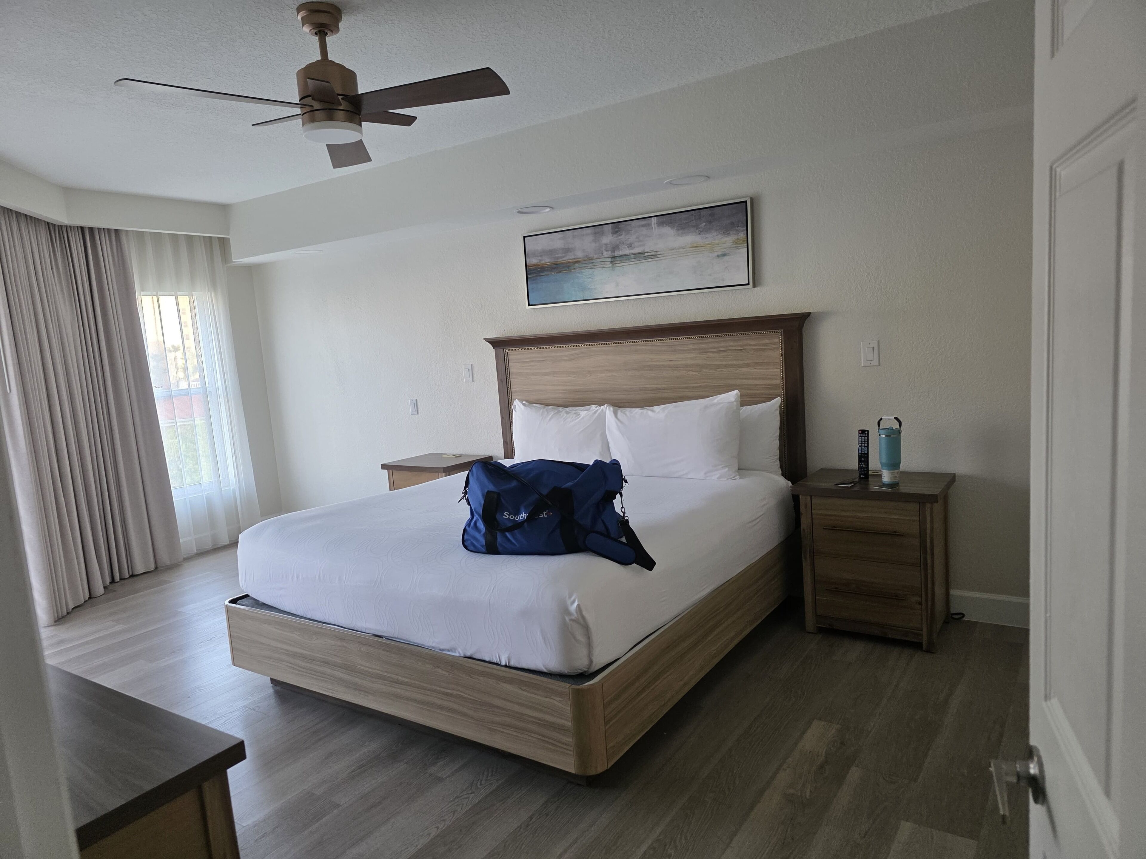 A hotel room with a bed, a nightstand, a ceiling fan, a painting, and a view of the outdoors.
