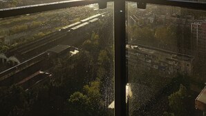 Un paisaje urbano con el sol poniéndose tras las colinas, visto desde un balcón con barandilla de vidrio.