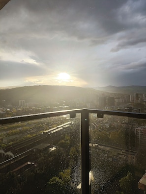 Stadtansicht mit untergehender Sonne hinter Hügeln, aufgenommen vom Balkon mit Glasgeländer.