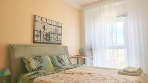 Un dormitorio con una cama, una ventana con cortinas translúcidas, un cuadro colgado en la pared y una mesita de noche con lámpara.