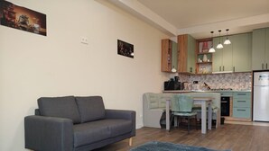 Una sala de estar con un sofá gris, un tapete azul, una zona de comedor con mesa y sillas, y una cocina con armarios verdes y un refrigerador blanco.