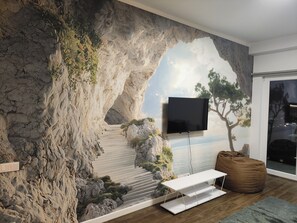 Uma sala de estar com um mural grande de uma paisagem rochosa, uma TV de tela plana na parede, uma mesa branca e uma poltrona de couro marrom.