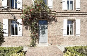 Un bâtiment en briques avec une porte centrale, des volets blancs et une vigne ornée de fleurs rouges.