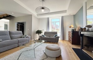 Un salon moderne avec un canapé gris, une table basse en verre, un pouf rond, une cheminée et une grande fenêtre avec des rideaux.
