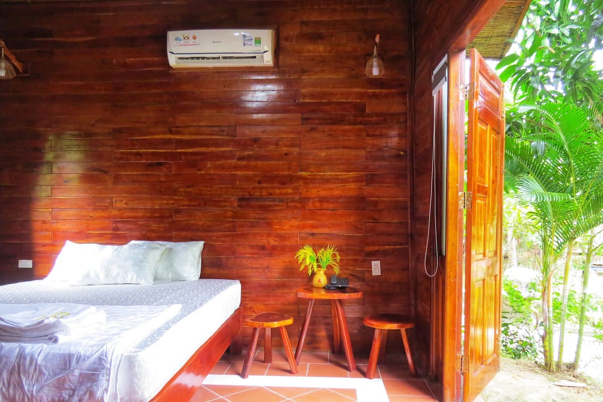 Una habitación de madera con una cama, una mesita con una planta y una puerta que da al exterior.