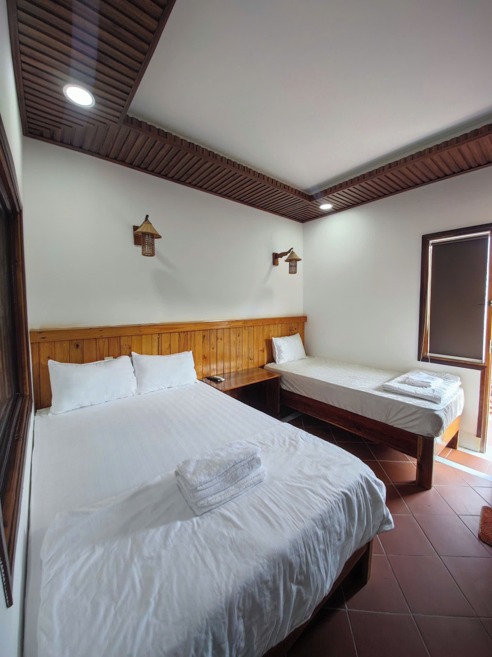 A hotel room with two beds, wooden headboard, wall-mounted lamps, and a window.