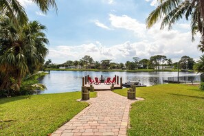 A pathway leading to a dock with red chairs and a fire pit by a calm lake, surrounded by palm trees and greenery. - Waterfront 3BR! Spacious Dock, King Bed, BBQ (Delray Beach)