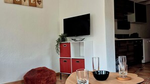 Una sala de estar con un televisor montado en la pared, un mueble rojo, una mesa de madera con vasos y un sillón de peluche rojo.