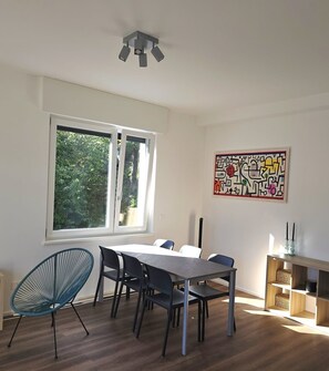 Een kamer met een eettafel, stoelen, een raam met uitzicht op groen, een plafondlamp en een wanddecoratie.