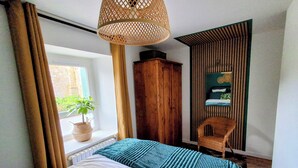Ett sovrum med en säng, ett fönster med gardiner, en krukväxt, en trägarderob, en stol och en hängande lampa.