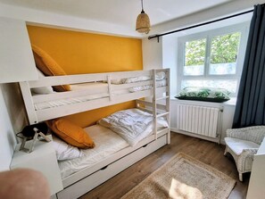 En køjeseng med hvidt sengetøj, en fletstol, en radiator og et vindue med udsigt til grønne omgivelser.