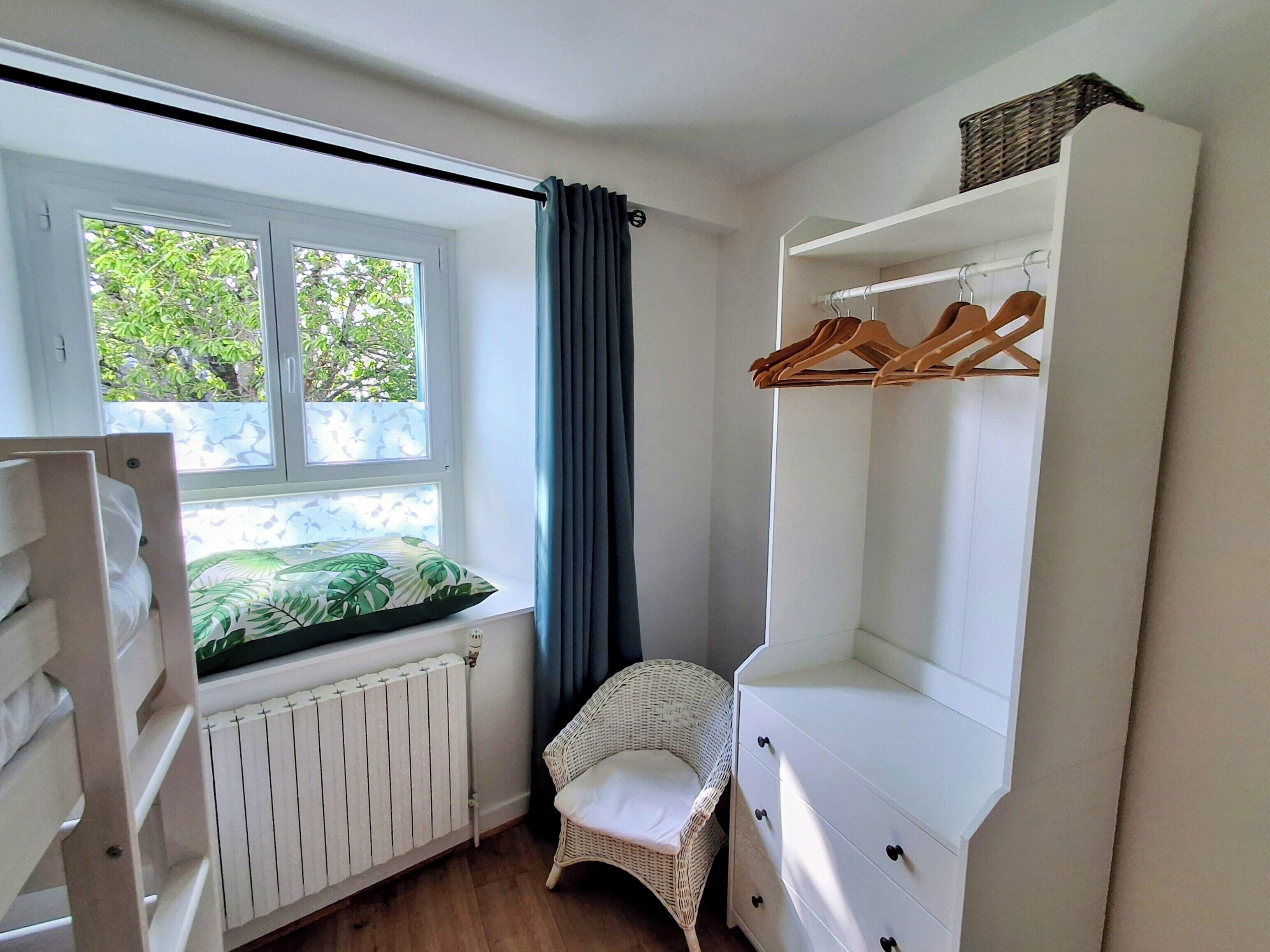 Ein Schlafzimmer mit Fenster, Heizkörper, weißem Schminktisch, Korbstuhl und Kleiderständer.