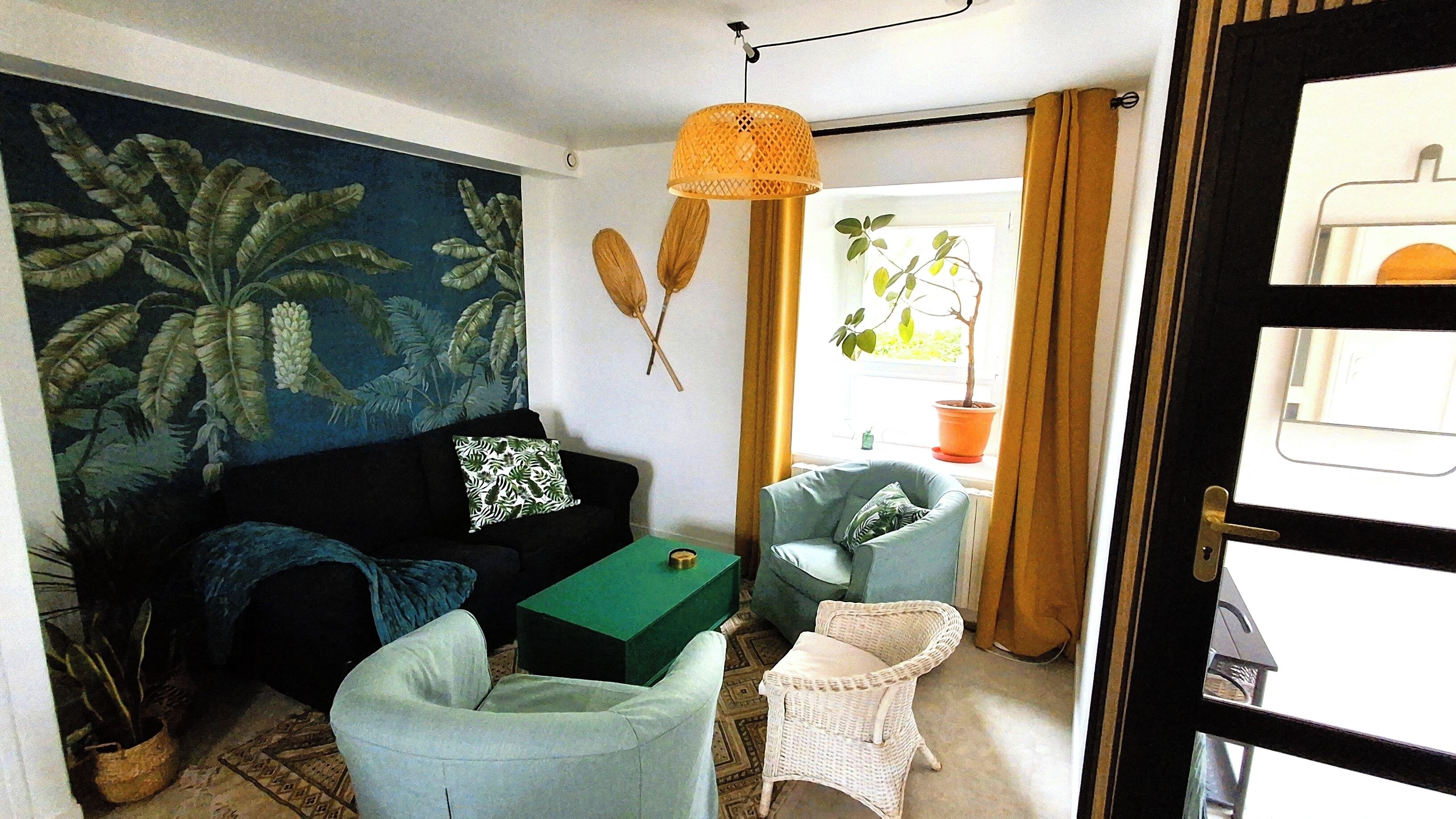 Un salón con un sofá oscuro, una mesa de centro verde, un sillón de mimbre y una pequeña silla blanca. Hay un cuadro en la pared, una lámpara colgante y una puerta que ofrece vistas al exterior.