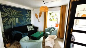 En stue med en mørk sofa, et grønt sofabord, en fletstol og en lille hvid stol. Der er et billede på væggen, en hængende lampe og en dør med udsigt ud.