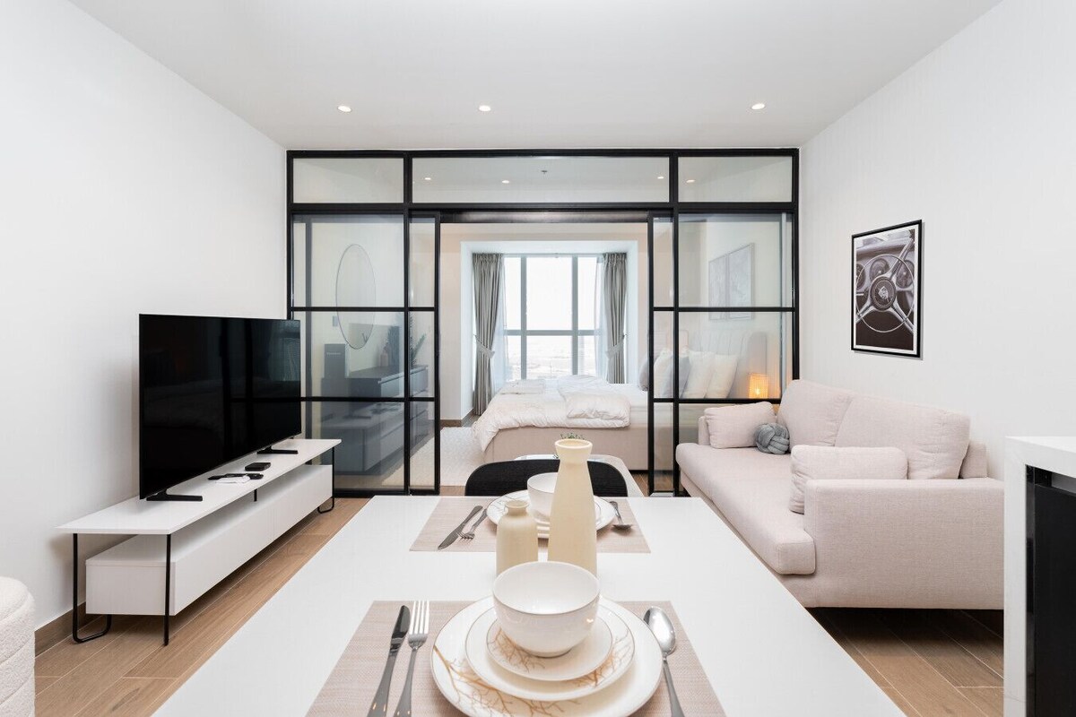 Et moderne hotelværelse med et fladskærms-tv, et spiseområde med et bord dækket til to personer, en sofa og et stort vindue med gardiner.