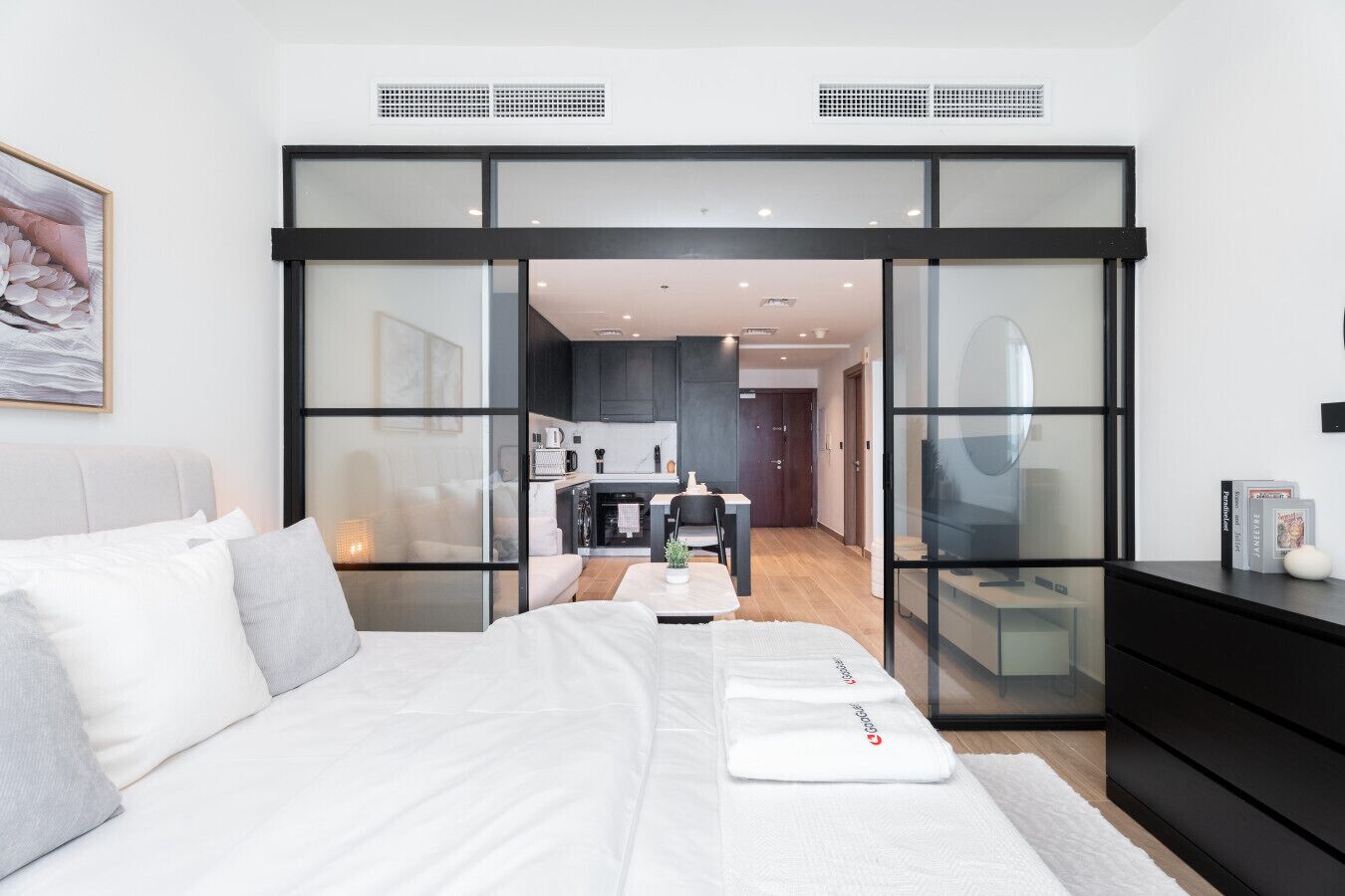 A modern bedroom with a large bed, a glass partition, a kitchen area, and a view into the living space.