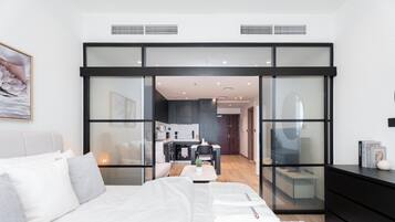 A modern bedroom with a large bed, a glass partition, a kitchen area, and a view into the living space.