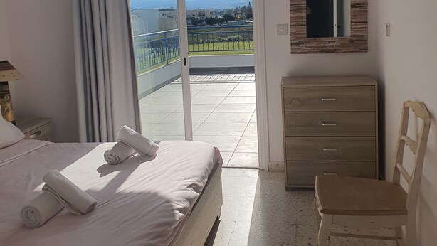 A bedroom with a bed, a dresser, a chair, a mirror, and a view of the outside. - Latchi Harbour 2-bedroom Penthouse (Poli Chrysochous)