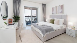A modern bedroom with a large bed, a desk, a chair, and a balcony with a city view. - Gala Guests - Princess Tower Suites (Dubai)