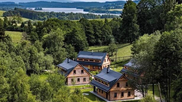 A wooden cabin complex with multiple buildings, surrounded by lush greenery and a lake in the background.