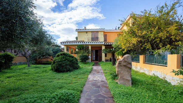 A stone pathway leading to a house with a tiled roof, surrounded by lush greenery and a stone wall. - BeLocations - Villa 50 meters from the sea (Solanas)
