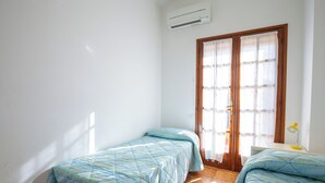 A room with two beds, an air conditioning unit, a wooden door, and a small desk with a lamp. - BeLocations - Villa 50 meters from the sea (Solanas)