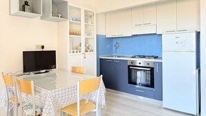 Cozinha moderna com armários brancos e azuis, fogão a gás, forno e geladeira embutida. Há uma mesa de jantar com cadeiras e uma TV na parede.