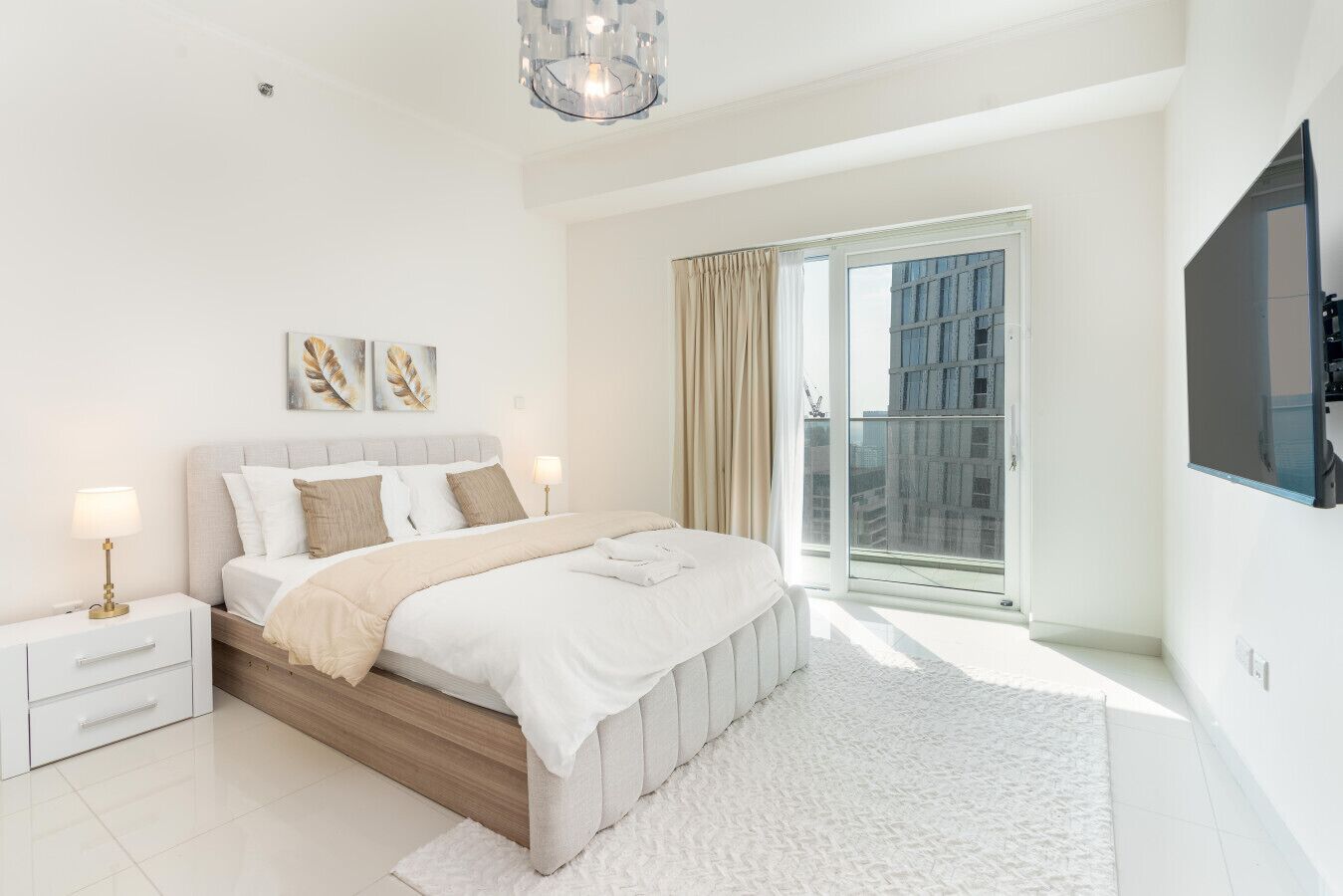 Une chambre moderne avec un lit, des tables de chevet, un lustre et une grande fenêtre donnant sur des immeubles.