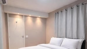 Una cama bien hecha con ropa blanca, una puerta cerrada y una pared de color claro con una cortina.