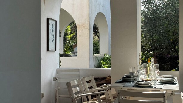 An outdoor dining area with a table set for two, surrounded by white chairs, under a covered structure with arches. - Cactus Cottage With pool and a stone's throw from the famous Blue Grotto! (Anacapri)