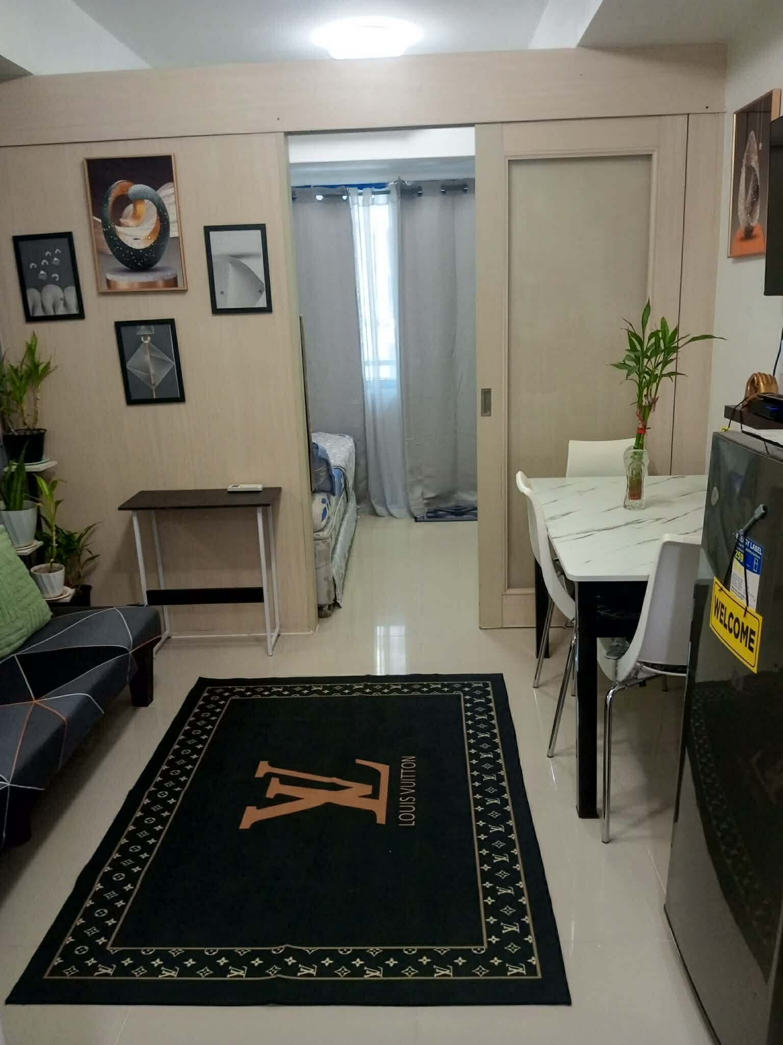 A living area with a black rug featuring a large 'LV' logo, a sofa, a dining table with chairs, a small table, and a potted plant.
