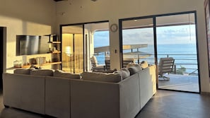 A living room with a sofa, TV, and a view of the sea through large glass doors.