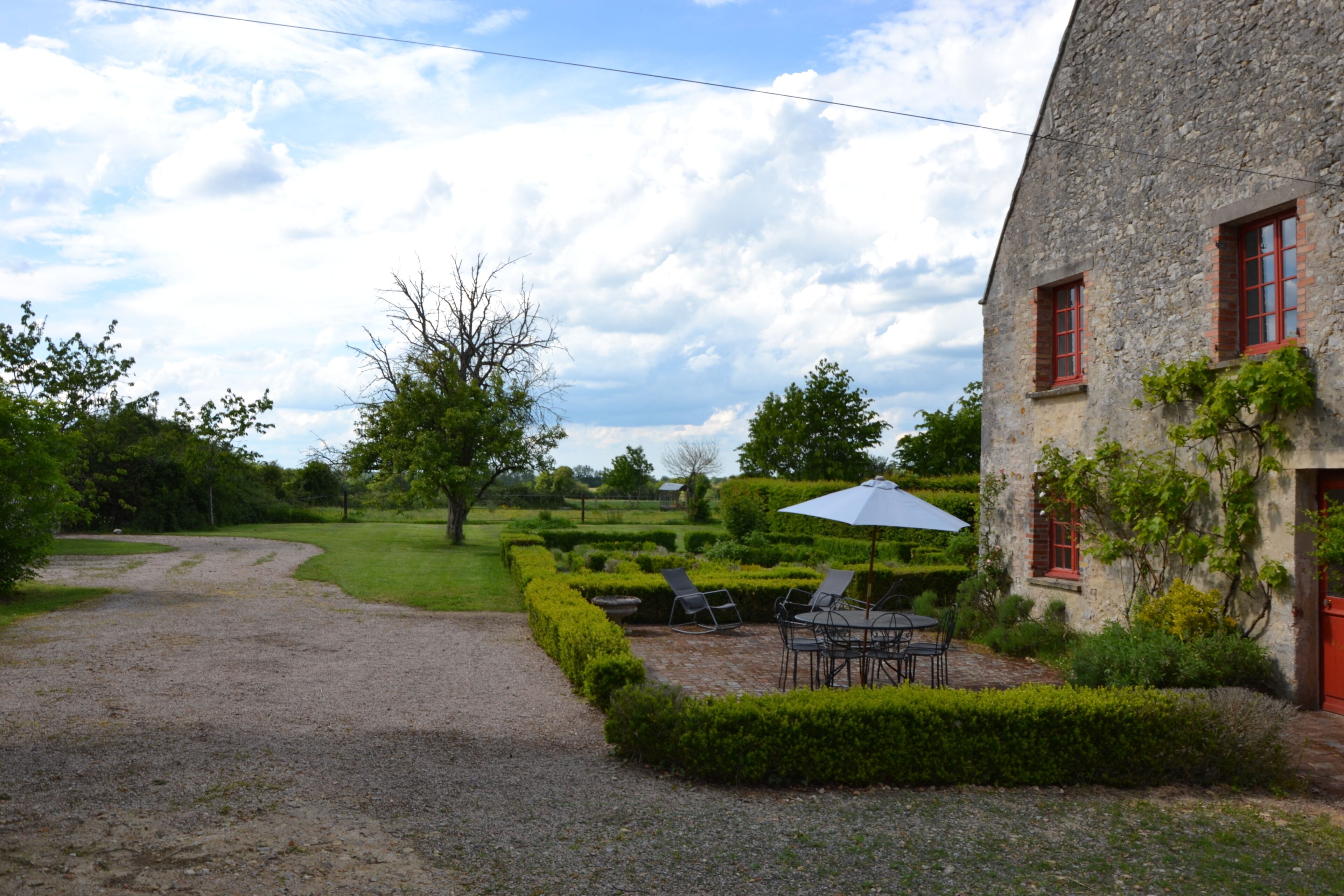 A stone building with red windows and a gravel path leading to a neatly trimmed garden with a table and chairs.