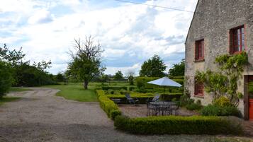 A stone building with red windows and a gravel path leading to a neatly trimmed garden with a table and chairs.