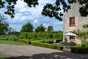 Un bâtiment en pierre avec une fenêtre rouge, un chemin de gravier, des haies soigneusement taillées et des sièges installés à l’extérieur.