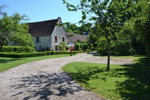 Une maison de campagne avec une allée en gravier, entourée de haies et d’arbres bien entretenus.