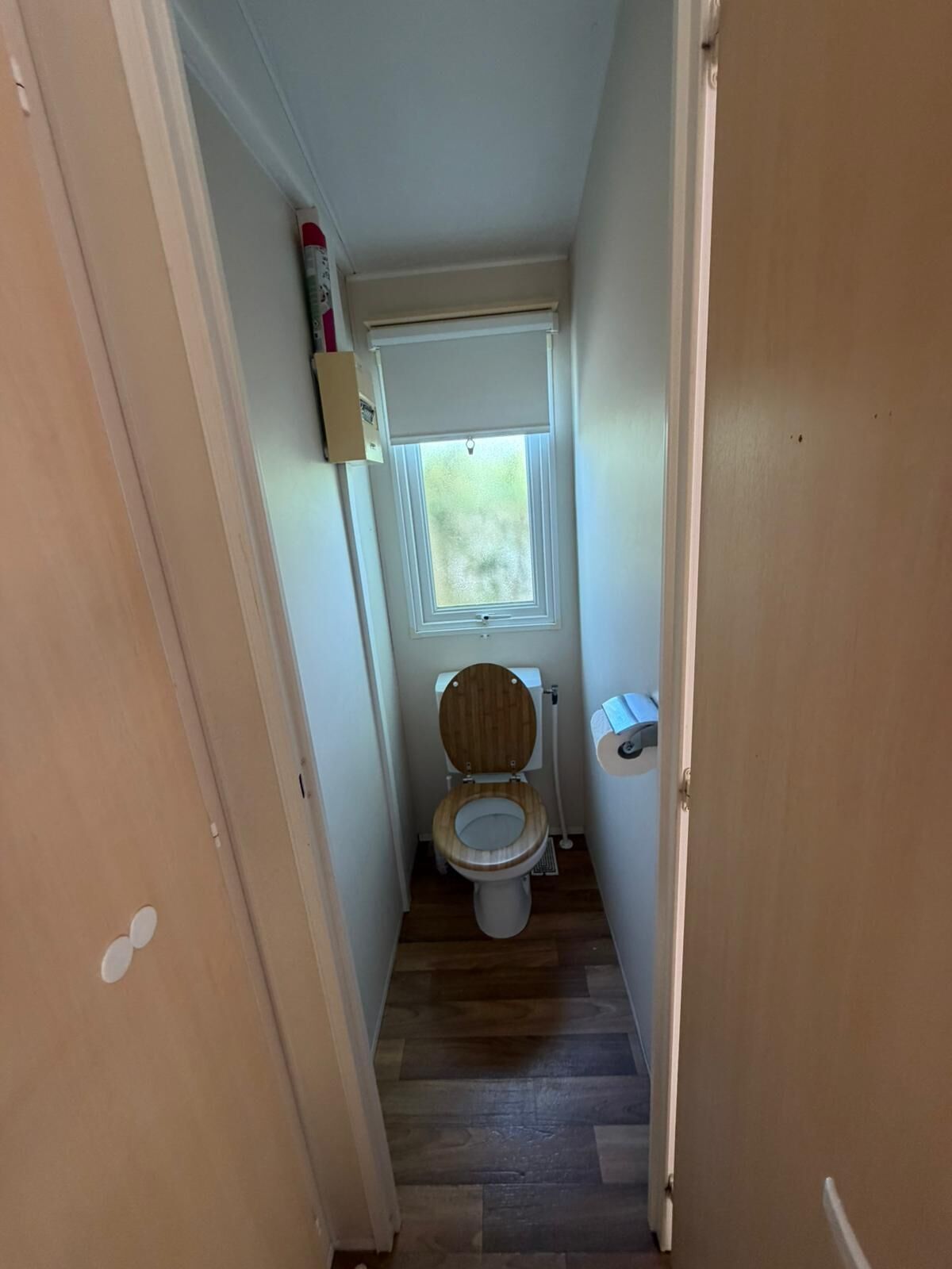 Un baño estrecho con inodoro de madera, una ventana sobre el inodoro y una puerta a la izquierda.