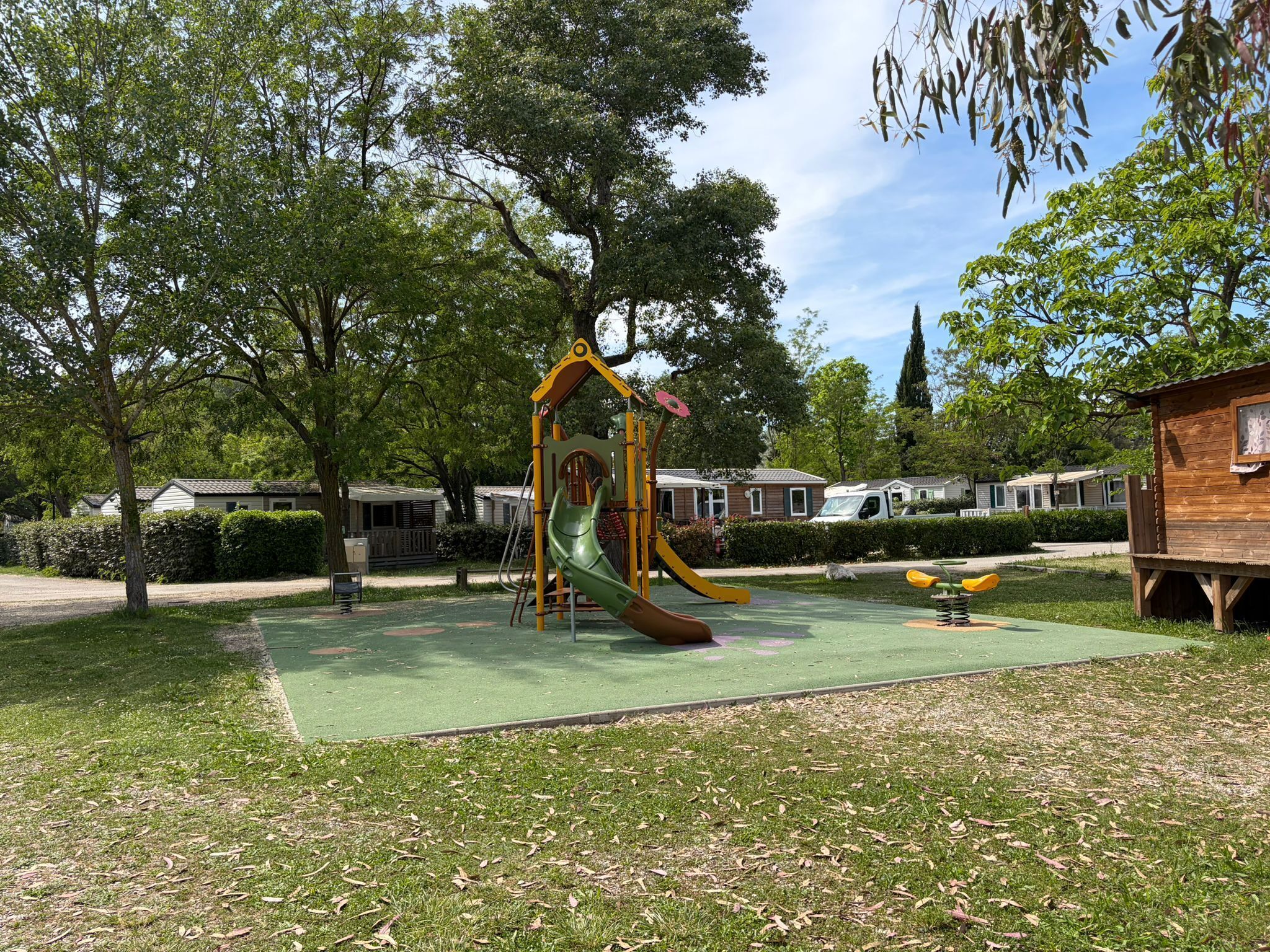 Une aire de jeux avec un toboggan et des balançoires, entourée d’arbres et d’une petite structure en bois.