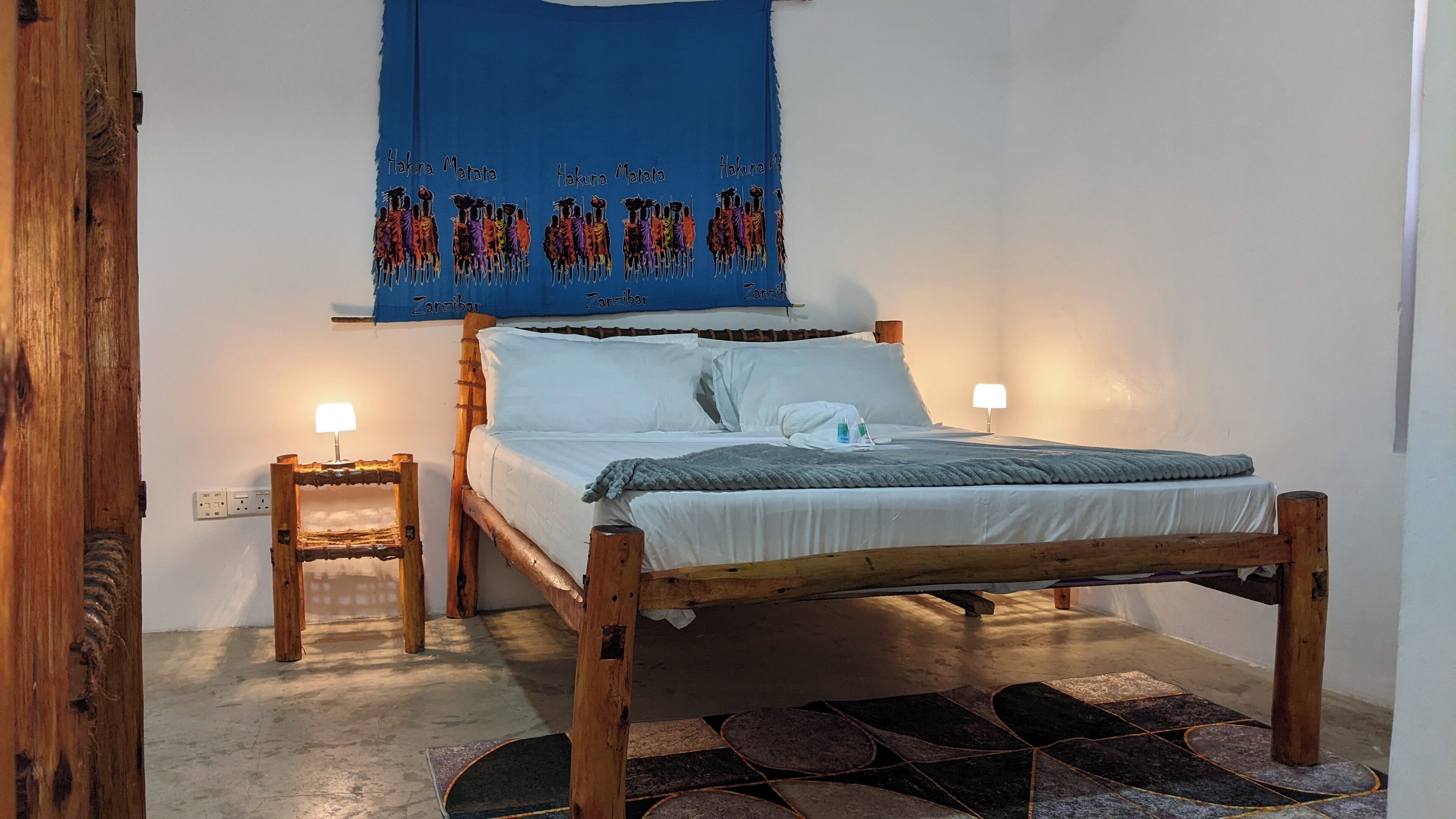 A room with a wooden bed, two bedside tables, a blue wall hanging, and a patterned rug.