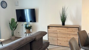 A modern living room with a flat-screen TV, a sofa, a dining area with chairs, a wooden cabinet, and a potted plant.