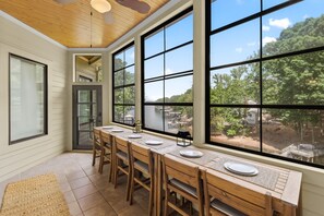A dining area with a long wooden table, chairs, and large windows offering a view of the outdoors.