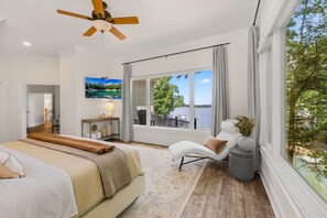 A bedroom with a bed, a chair, a desk, a TV, and a large window with a view of a body of water.