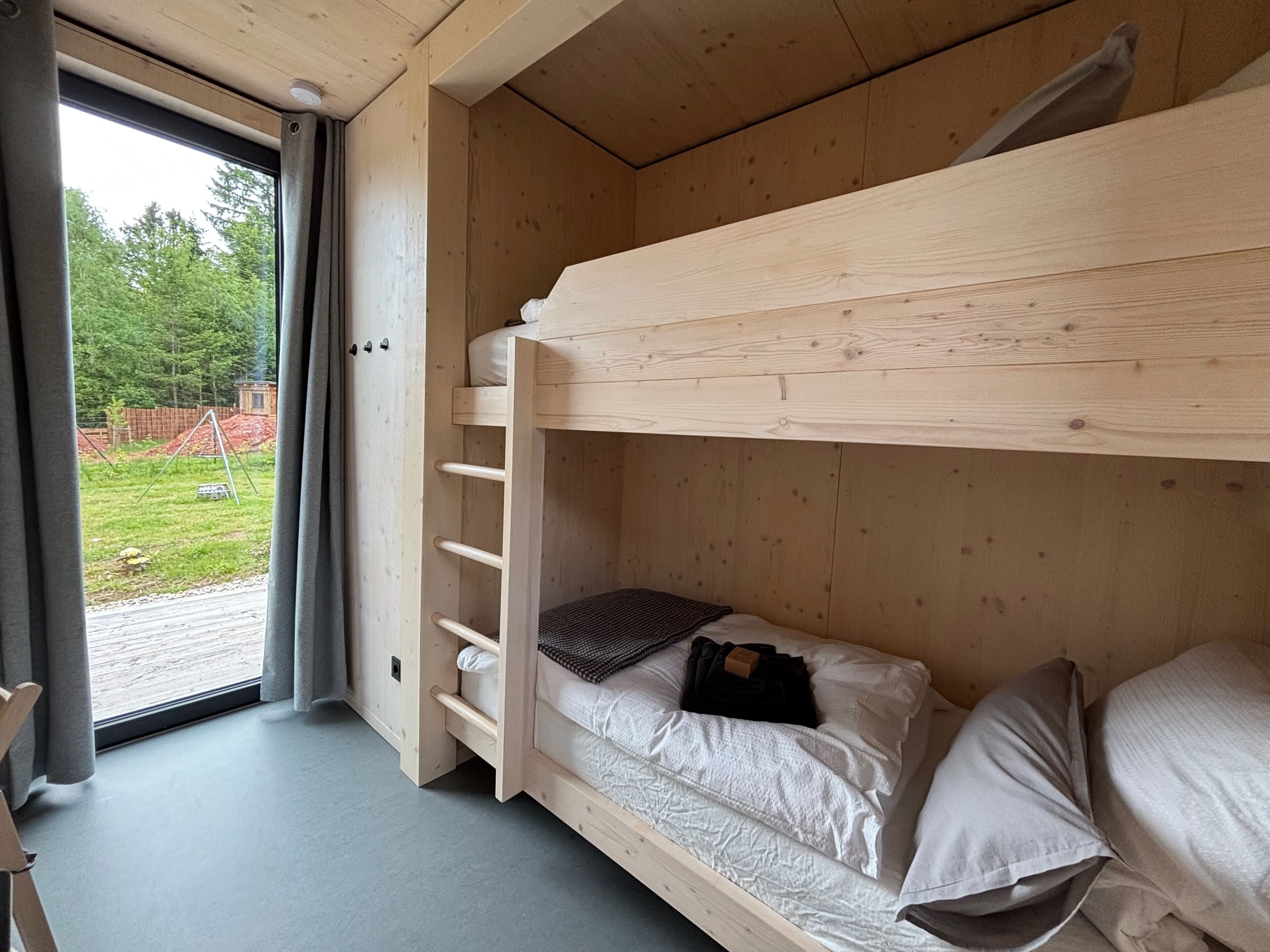 A bunk bed with a wooden ladder, a window with curtains, and a view of a green landscape.
