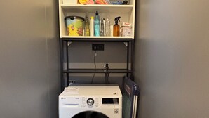 A compact laundry area with a washing machine, shelves with cleaning supplies, and a power outlet.