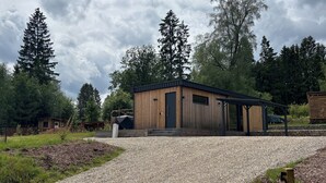 A modern wooden cabin with a gravel driveway, surrounded by trees.