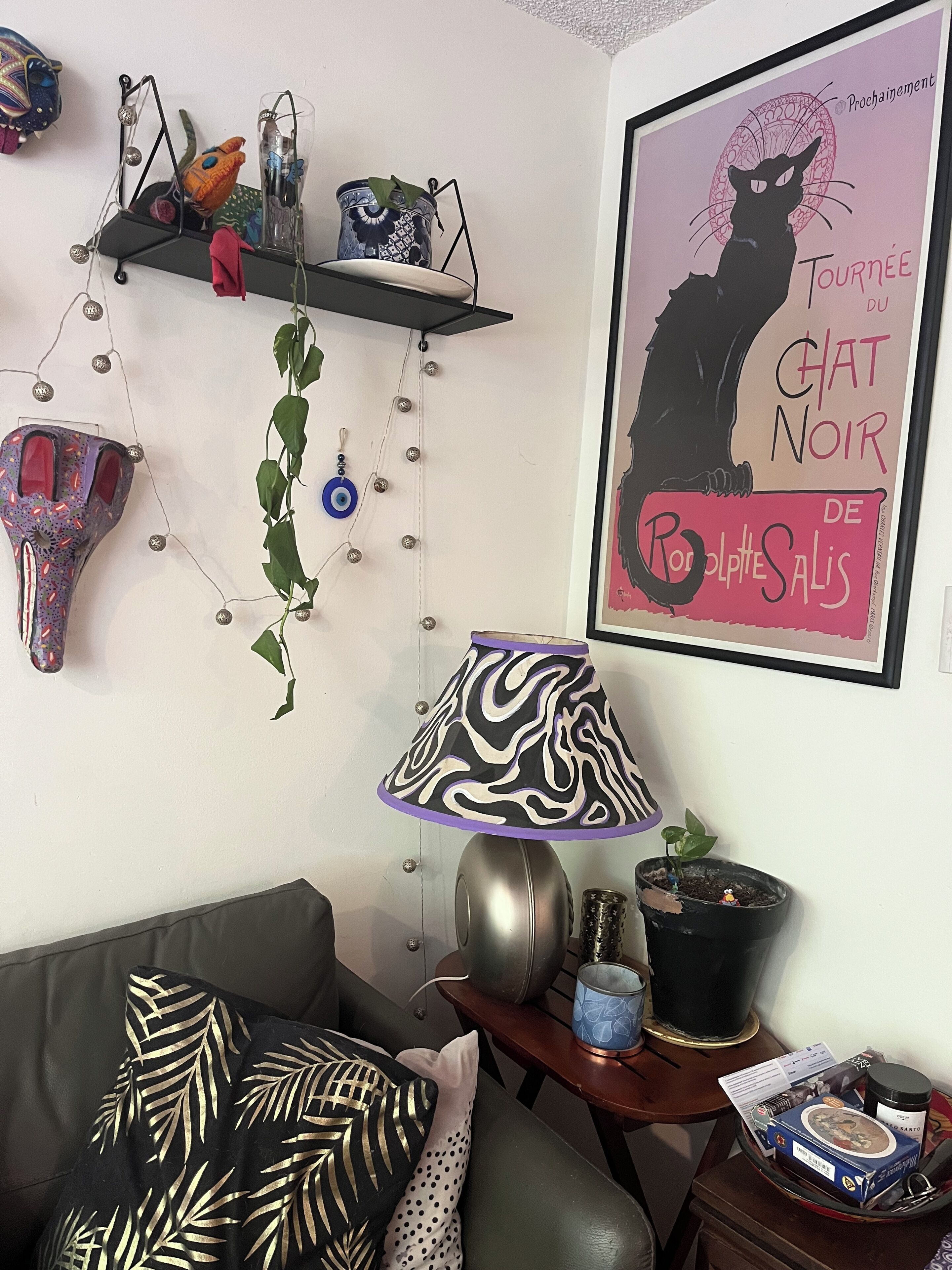 A room with a sofa, a lamp, a framed poster, a shelf with decorative items, and a small table with a plant and a box.