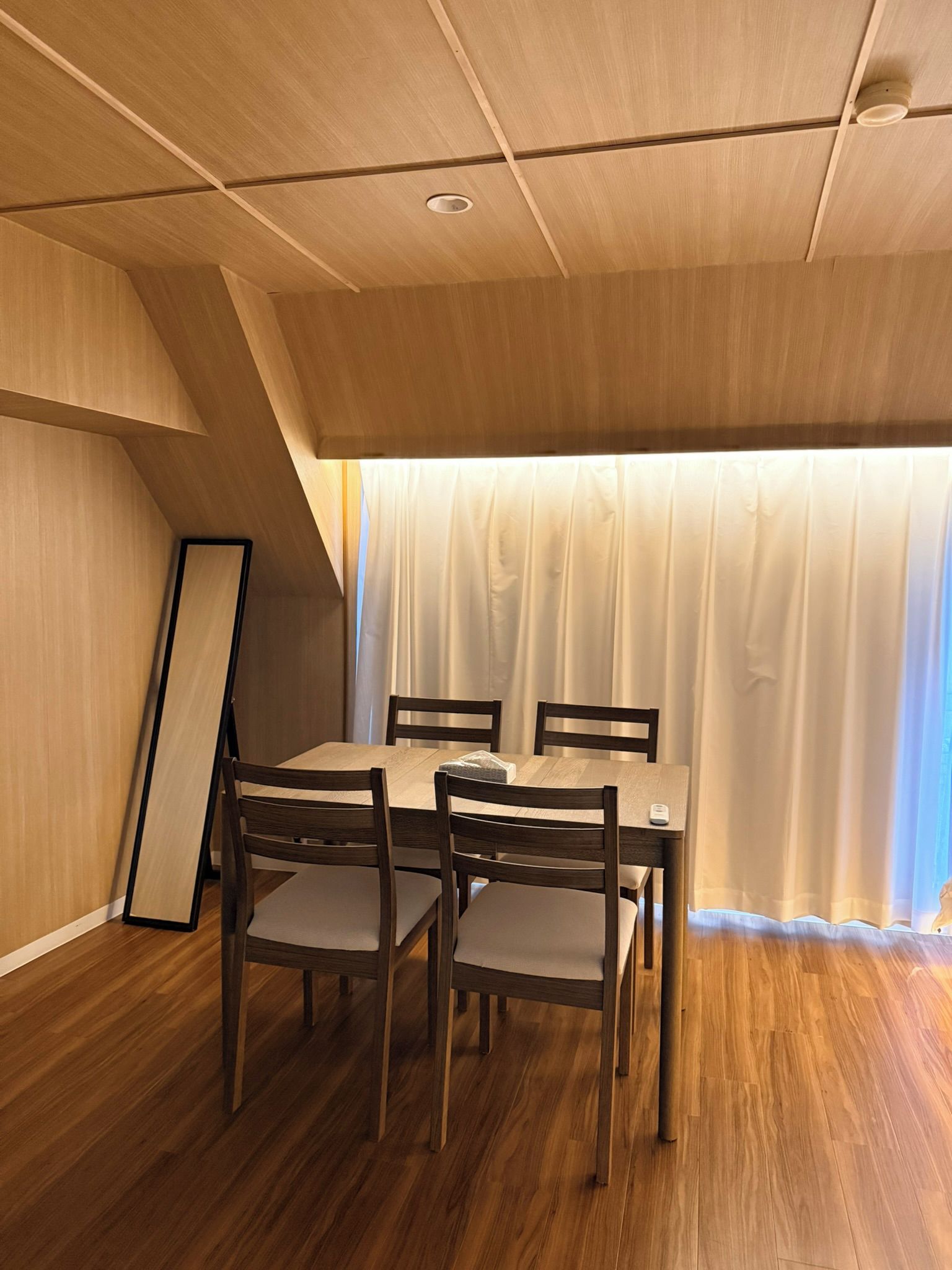 A dining area with a wooden table and chairs, a ceiling with recessed lighting, and a view through a window with curtains.